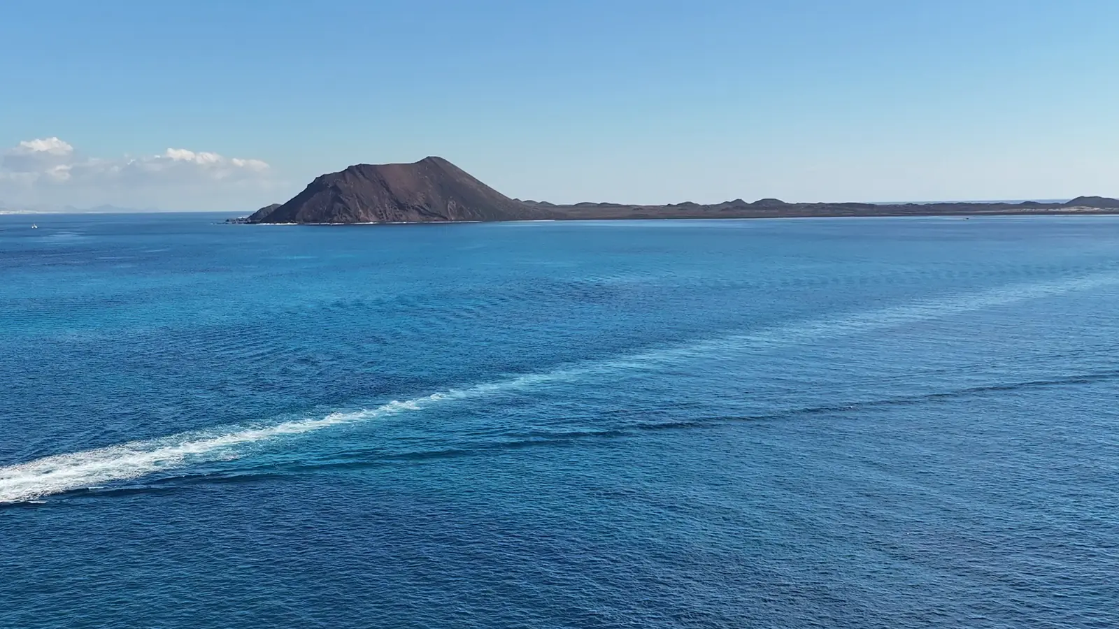 Vista aérea de Isla de Lobos desde el mar: el cono volcánico y la costa baja en el horizonte, con la estela blanca de una embarcación sobre el agua azul. Fotografía de Grupo Oby