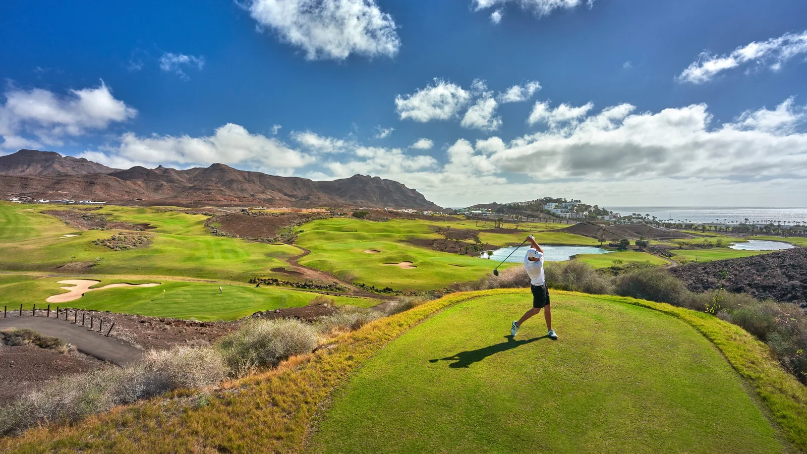 Golfista en el tee de salida de Playitas Golf, con las calles del campo, dos lagos, montañas de tierra rojiza y el mar al fondo
